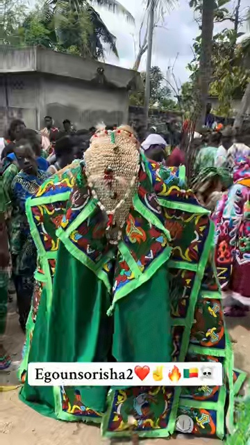 Egungun au costume vert brodé de motifs colorés, masque couvert de cauris, entouré de la foule